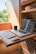A compact workspace within a small wooden room, possibly a van or camper, featuring a wooden table with a laptop and a keyboard. The table is foldable and the room also contains some shelves with items like bowls and plants. A mug is placed near the laptop, which displays a website. Natural light enters the space, creating a warm and cozy atmosphere.