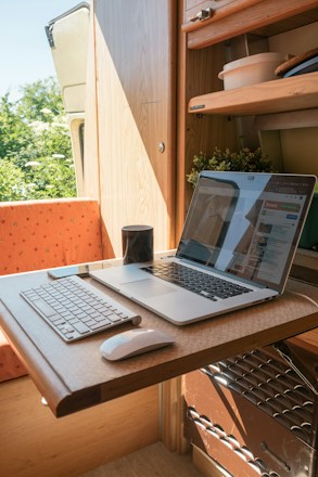 A compact workspace within a small wooden room, possibly a van or camper, featuring a wooden table with a laptop and a keyboard. The table is foldable and the room also contains some shelves with items like bowls and plants. A mug is placed near the laptop, which displays a website. Natural light enters the space, creating a warm and cozy atmosphere.