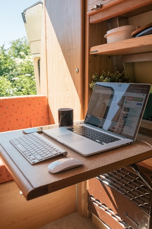 A compact workspace within a small wooden room, possibly a van or camper, featuring a wooden table with a laptop and a keyboard. The table is foldable and the room also contains some shelves with items like bowls and plants. A mug is placed near the laptop, which displays a website. Natural light enters the space, creating a warm and cozy atmosphere.
