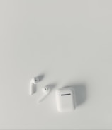 A pair of white wireless earbuds and a matching charging case are placed on a smooth, light-colored surface. The earbuds have a sleek and minimalist design, with one of them slightly tilted.