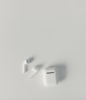 A pair of white wireless earbuds and a matching charging case are placed on a smooth, light-colored surface. The earbuds have a sleek and minimalist design, with one of them slightly tilted.