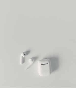 A pair of white wireless earbuds and a matching charging case are placed on a smooth, light-colored surface. The earbuds have a sleek and minimalist design, with one of them slightly tilted.