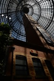 A large architectural dome made of metal and glass frames the top of a brick building, with geometric patterns forming concentric circles towards the center. The brick building features tall, arched windows with bars, and a vintage-style sign mounted on the wall. Shadows play across the brickwork, adding dramatic contrast.