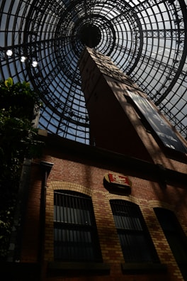 A large architectural dome made of metal and glass frames the top of a brick building, with geometric patterns forming concentric circles towards the center. The brick building features tall, arched windows with bars, and a vintage-style sign mounted on the wall. Shadows play across the brickwork, adding dramatic contrast.