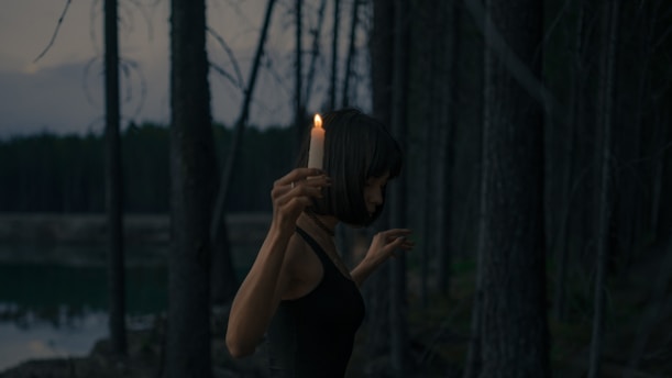 A shadowy figure holding a flickering candle in a misty forest at twilight.