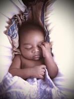 A sleeping baby rests peacefully, with one hand gently touching the face. The baby is partially covered with a patterned blanket, featuring shades of blue and intricate designs. The overall setting appears calm and intimate, highlighting the baby's serene expression.