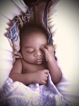 A sleeping baby rests peacefully, with one hand gently touching the face. The baby is partially covered with a patterned blanket, featuring shades of blue and intricate designs. The overall setting appears calm and intimate, highlighting the baby's serene expression.