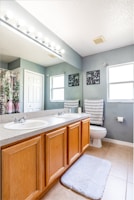 A modern bathroom featuring a double sink vanity with wooden cabinets, a large mirror above, and bright lighting. The space includes a toilet, a patterned towel, a soft floor mat, and decorative wall hangings. The walls are painted a light gray, complementing the tiled floor.