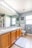 A modern bathroom featuring a double sink vanity with wooden cabinets, a large mirror above, and bright lighting. The space includes a toilet, a patterned towel, a soft floor mat, and decorative wall hangings. The walls are painted a light gray, complementing the tiled floor.