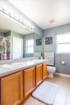 A modern bathroom featuring a double sink vanity with wooden cabinets, a large mirror above, and bright lighting. The space includes a toilet, a patterned towel, a soft floor mat, and decorative wall hangings. The walls are painted a light gray, complementing the tiled floor.