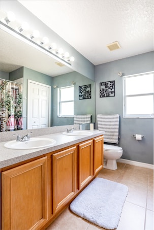 A modern bathroom featuring a double sink vanity with wooden cabinets, a large mirror above, and bright lighting. The space includes a toilet, a patterned towel, a soft floor mat, and decorative wall hangings. The walls are painted a light gray, complementing the tiled floor.