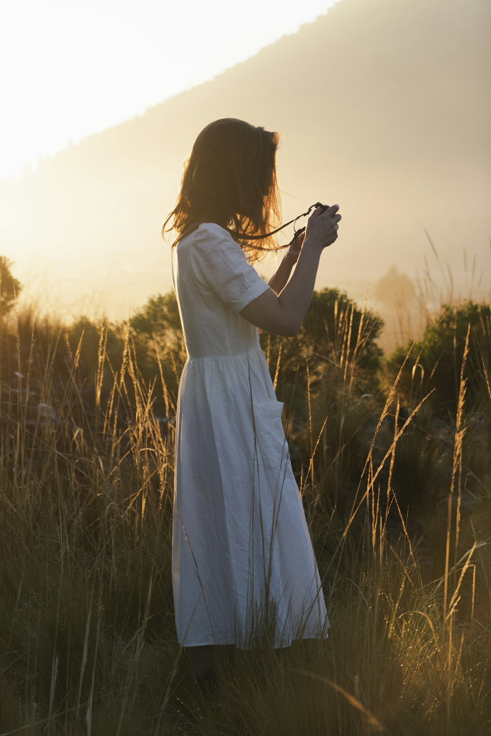 Frau im weißen Kleid mit Kamera steht während des Sonnenuntergangs auf grünem Grasfeld