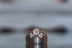 A close-up view of a luxurious diamond engagement ring set on top of a wedding band. The central diamond is a large, square cut, flanked by smaller round diamonds set into the gold band. The background is blurred, emphasizing the elegant details and sparkle of the jewelry.