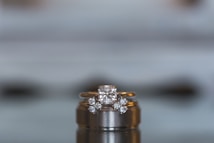 A close-up view of a luxurious diamond engagement ring set on top of a wedding band. The central diamond is a large, square cut, flanked by smaller round diamonds set into the gold band. The background is blurred, emphasizing the elegant details and sparkle of the jewelry.