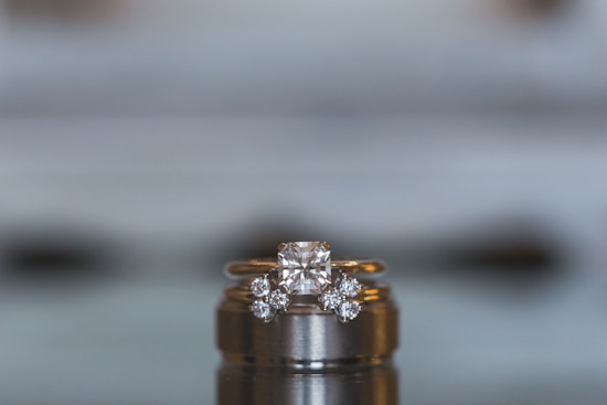 A close-up view of a luxurious diamond engagement ring set on top of a wedding band. The central diamond is a large, square cut, flanked by smaller round diamonds set into the gold band. The background is blurred, emphasizing the elegant details and sparkle of the jewelry.