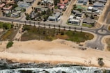 An aerial view of a coastal residential area featuring houses with landscaped gardens separated from the beach by a road lined with palm trees. The sandy beach meets the ocean with visible waves approaching the shore, and a few cars are parked near a roundabout.