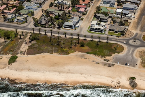 An aerial view of a coastal residential area featuring houses with landscaped gardens separated from the beach by a road lined with palm trees. The sandy beach meets the ocean with visible waves approaching the shore, and a few cars are parked near a roundabout.
