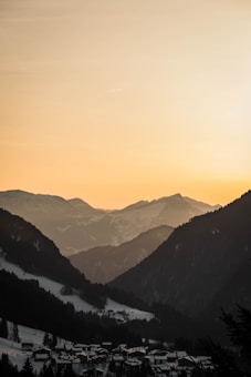 A tranquil mountainous landscape at sunset, with silhouettes of jagged peaks against a warm golden sky. Snow-covered hills and a quaint village rest in the valley below, creating a serene and peaceful scene.