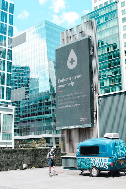 A large digital billboard in an urban setting displays a message encouraging blood donations, featuring an icon of a droplet with a plus sign. Surrounding the billboard are modern glass office buildings reflecting the sky. In the foreground, a person stands on rollerblades, looking at a phone next to a colorful blue trailer with branding on the side.