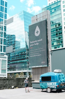 A large digital billboard in an urban setting displays a message encouraging blood donations, featuring an icon of a droplet with a plus sign. Surrounding the billboard are modern glass office buildings reflecting the sky. In the foreground, a person stands on rollerblades, looking at a phone next to a colorful blue trailer with branding on the side.
