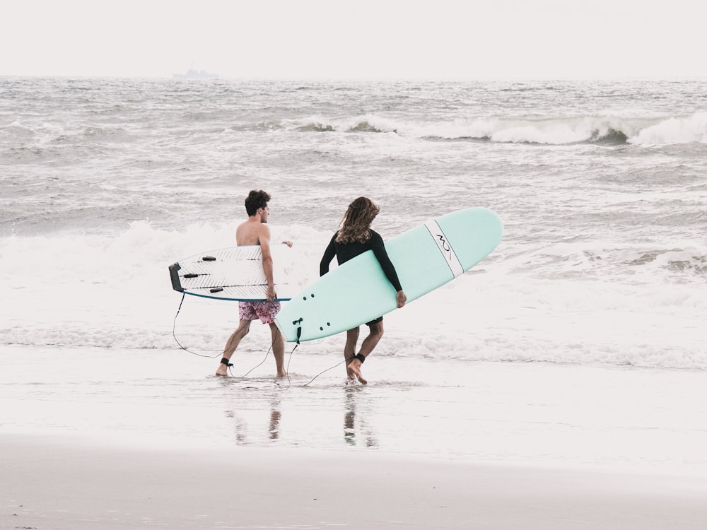 2 femmes tenant une planche de surf bleue et blanche marchant sur la plage pendant la journée
