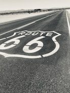A long road stretches into the distance, marked with a large Route 66 emblem on the asphalt. Surrounding the road is a barren desert landscape with distant mountains on the horizon, under a clear sky.
