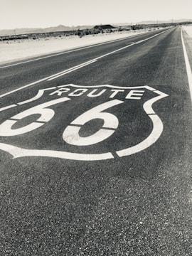 A long road stretches into the distance, marked with a large Route 66 emblem on the asphalt. Surrounding the road is a barren desert landscape with distant mountains on the horizon, under a clear sky.