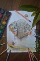 A detailed drawing of an elephant with colorful butterfly-inspired ears. The paper is surrounded by art supplies such as colored pencils, an eraser, and a watercolor palette. Next to the drawing, green leaves and a small flower add a natural element.