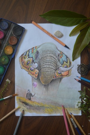 A detailed drawing of an elephant with colorful butterfly-inspired ears. The paper is surrounded by art supplies such as colored pencils, an eraser, and a watercolor palette. Next to the drawing, green leaves and a small flower add a natural element.