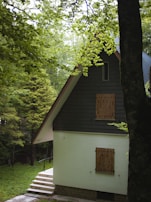 A cozy, secluded house with a steep A-frame roof surrounded by lush green trees. The cottage has a light-colored exterior with wooden shutters on the windows. A small set of steps leads up to the entrance, and the foliage around the house creates a tranquil and inviting atmosphere.