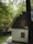 A cozy, secluded house with a steep A-frame roof surrounded by lush green trees. The cottage has a light-colored exterior with wooden shutters on the windows. A small set of steps leads up to the entrance, and the foliage around the house creates a tranquil and inviting atmosphere.