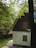 A cozy, secluded house with a steep A-frame roof surrounded by lush green trees. The cottage has a light-colored exterior with wooden shutters on the windows. A small set of steps leads up to the entrance, and the foliage around the house creates a tranquil and inviting atmosphere.