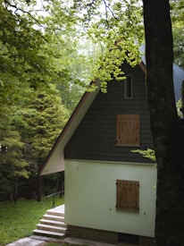 A cozy, secluded house with a steep A-frame roof surrounded by lush green trees. The cottage has a light-colored exterior with wooden shutters on the windows. A small set of steps leads up to the entrance, and the foliage around the house creates a tranquil and inviting atmosphere.