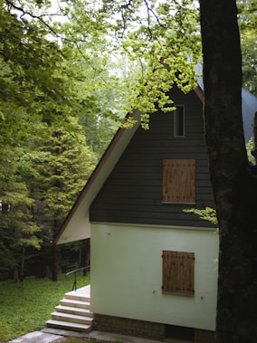 A cozy, secluded house with a steep A-frame roof surrounded by lush green trees. The cottage has a light-colored exterior with wooden shutters on the windows. A small set of steps leads up to the entrance, and the foliage around the house creates a tranquil and inviting atmosphere.