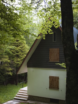 A cozy, secluded house with a steep A-frame roof surrounded by lush green trees. The cottage has a light-colored exterior with wooden shutters on the windows. A small set of steps leads up to the entrance, and the foliage around the house creates a tranquil and inviting atmosphere.