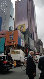 A busy urban street corner with high-rise buildings covered in large, colorful advertisements and billboards. Several vehicles, including a taxi, SUV, and a truck, are waiting at the intersection. A cyclist rides by while a man in a suit stands smiling in the foreground. The sky is overcast, adding a dull tone to the scene.