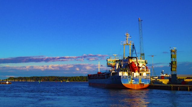 A large cargo ship docked at a port on a calm body of water. The sky is clear with a vibrant blue hue, and the sun casts warm lighting on the structures at the dock. A lush tree line is visible on the opposite shore, enhancing the serene and peaceful setting.