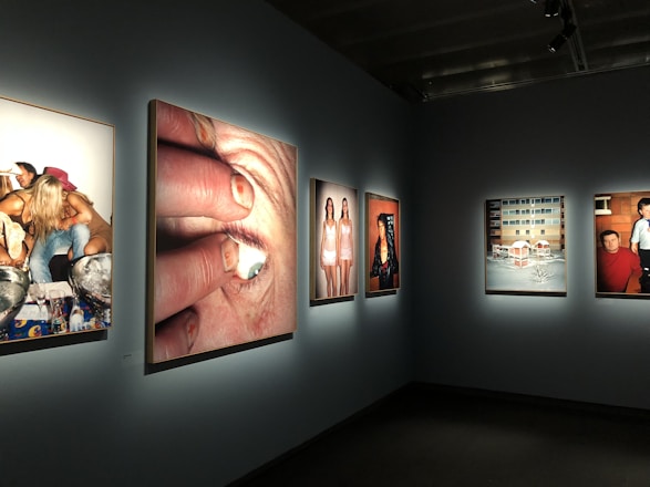 A gallery room is displaying several framed photographs on a wall. The images exhibit various scenes, including a close-up of a person's eye being held open, a group of people partying, two women posing in matching outfits, a snowy residential building scene, and a casual snapshot of two individuals indoors.