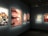 A gallery room is displaying several framed photographs on a wall. The images exhibit various scenes, including a close-up of a person's eye being held open, a group of people partying, two women posing in matching outfits, a snowy residential building scene, and a casual snapshot of two individuals indoors.