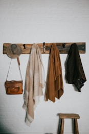A rustic wooden coat rack mounted on a white brick wall holds a grey hat, a brown leather bag, and three woolen scarves or shawls in muted beige and brown tones. Below the rack, a small wooden stool sits on the floor.