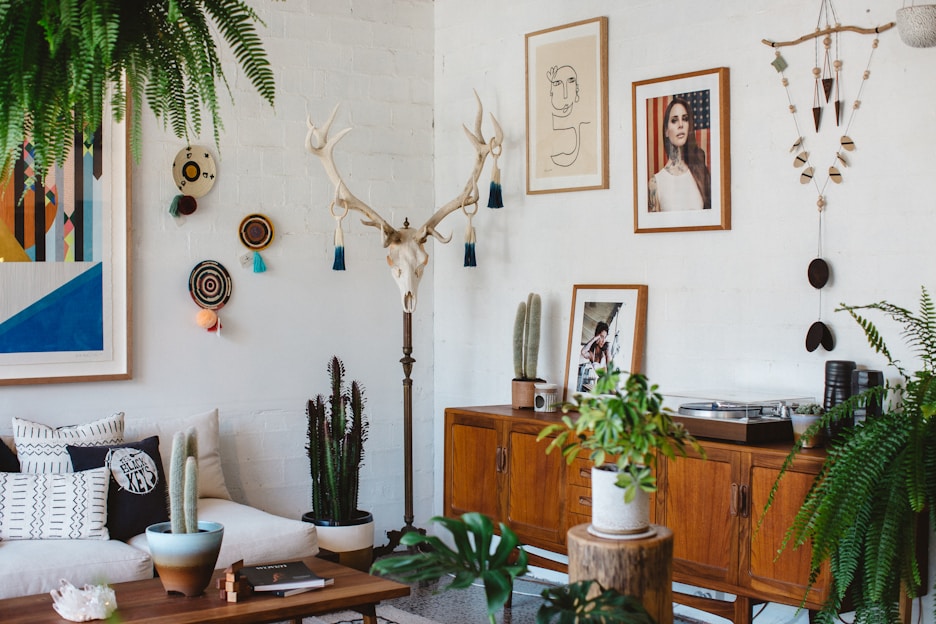 A living room with a bohemian style, featuring a white brick wall decorated with eclectic art pieces and a mounted antler. There is a wooden sideboard displaying cacti and a turntable. A mix of cushions is arranged on a light-colored couch, and a variety of potted plants are spread throughout, including ferns and succulents.