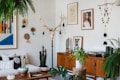 A living room with a bohemian style, featuring a white brick wall decorated with eclectic art pieces and a mounted antler. There is a wooden sideboard displaying cacti and a turntable. A mix of cushions is arranged on a light-colored couch, and a variety of potted plants are spread throughout, including ferns and succulents.