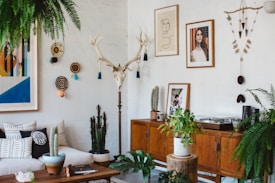 A living room with a bohemian style, featuring a white brick wall decorated with eclectic art pieces and a mounted antler. There is a wooden sideboard displaying cacti and a turntable. A mix of cushions is arranged on a light-colored couch, and a variety of potted plants are spread throughout, including ferns and succulents.
