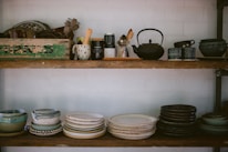 Two wooden shelves filled with various kitchenware and pottery, including stacks of bowls and plates in earthy tones. The top shelf features a variety of kitchen utensils, a teapot, and a rustic green box with text on it. The arrangement has a cozy, vintage feel.