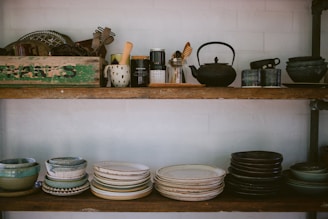 Two wooden shelves filled with various kitchenware and pottery, including stacks of bowls and plates in earthy tones. The top shelf features a variety of kitchen utensils, a teapot, and a rustic green box with text on it. The arrangement has a cozy, vintage feel.