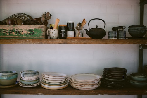 An organized kitchen shelf featuring earthy-toned ceramic jars, woven baskets, and neatly stacked plates.