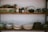 Two wooden shelves filled with various kitchenware and pottery, including stacks of bowls and plates in earthy tones. The top shelf features a variety of kitchen utensils, a teapot, and a rustic green box with text on it. The arrangement has a cozy, vintage feel.