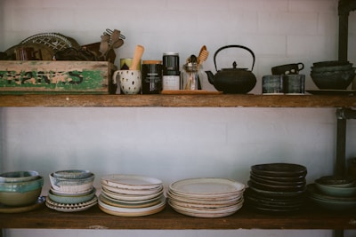 Two wooden shelves filled with various kitchenware and pottery, including stacks of bowls and plates in earthy tones. The top shelf features a variety of kitchen utensils, a teapot, and a rustic green box with text on it. The arrangement has a cozy, vintage feel.