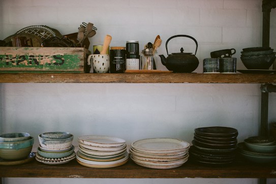 Two wooden shelves filled with various kitchenware and pottery, including stacks of bowls and plates in earthy tones. The top shelf features a variety of kitchen utensils, a teapot, and a rustic green box with text on it. The arrangement has a cozy, vintage feel.