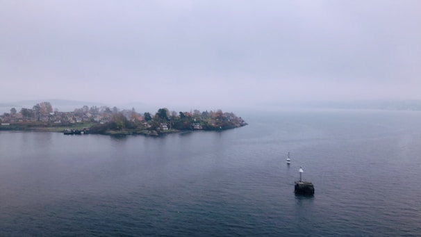 A calm and expansive body of water under a cloudy sky, with a small island that features several houses surrounded by trees. Two buoys are visible in the water, and the shoreline of the island appears lush with greenery.